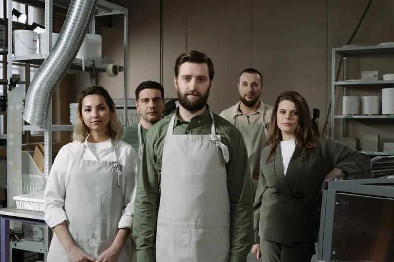 Five people in aprons standing together in a pottery studio with shelves and equipment behind them.