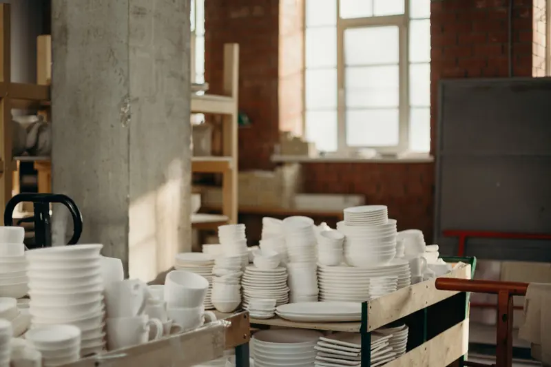 Stacks of handmade white bowls, plates and mugs on shelves in a pottery workshop, the kind of tableware a clay body selector helps you plan.