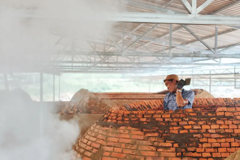A worker standing on a large brick kiln with steam rising around it.