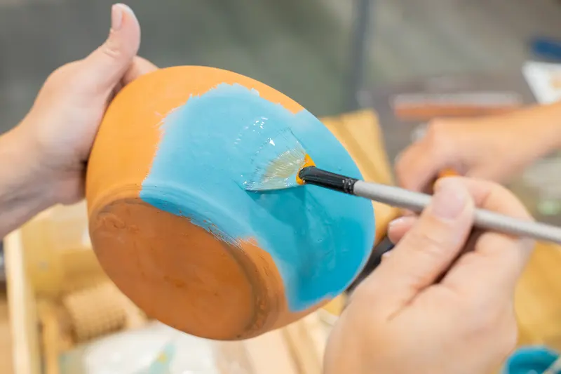 Hands brushing blue glaze onto a ceramic bowl, applying coats as the AMACO glaze combinations flyers describe.
