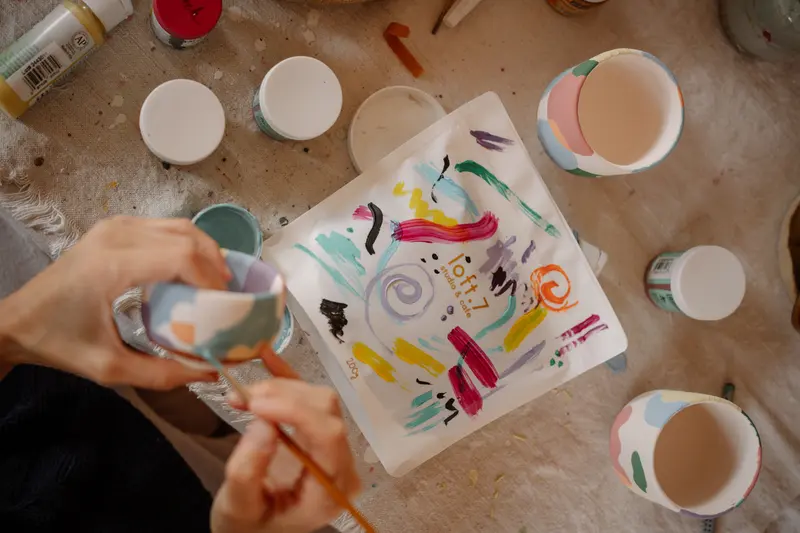 Hands painting colourful designs on pottery in a workshop, using underglaze colours like Speedball underglaze.