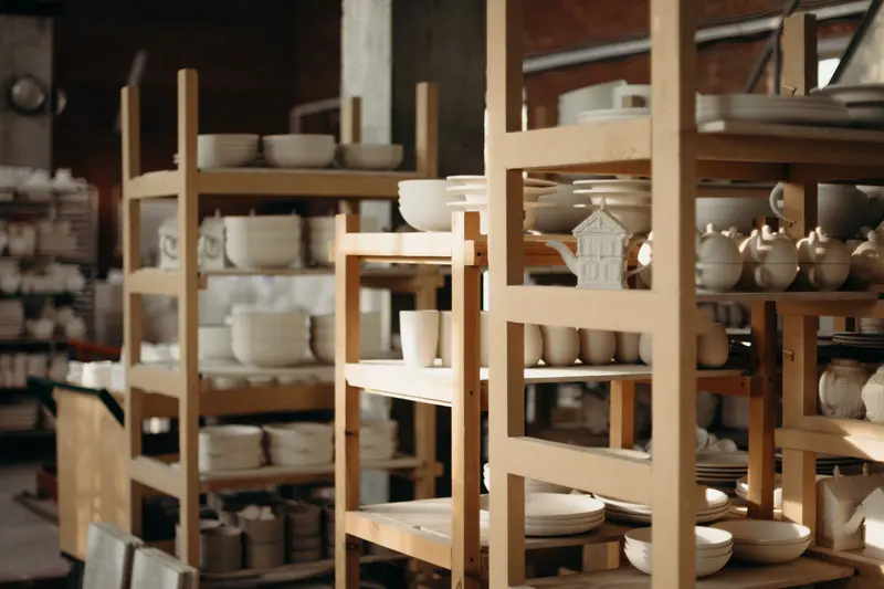 Wooden shelves full of handmade white bowls, mugs and teapots in a busy pottery workshop.