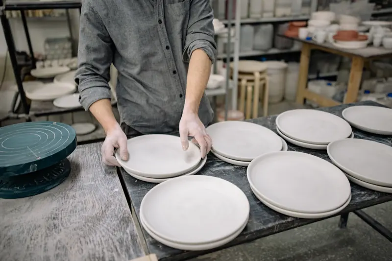 A potter arranging freshly made plates on a studio table ahead of firing, where kiln electricity cost depends on how full the load is.