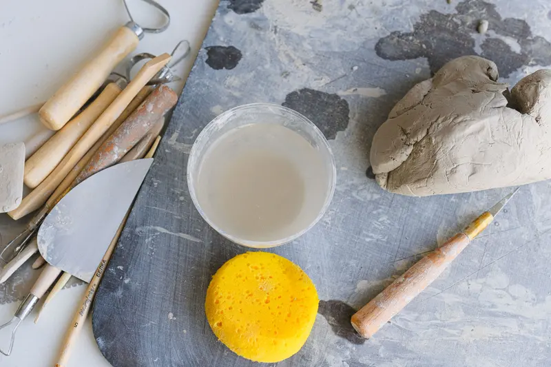 Pottery tools, a yellow sponge, a cup of water and a lump of clay on a grey board.