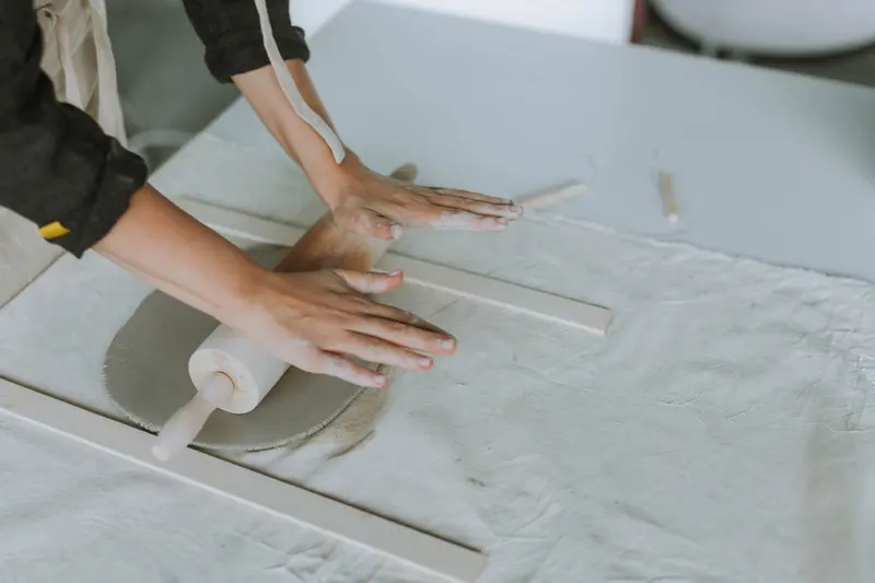 Hands rolling a clay slab with a rolling pin between two guide sticks.