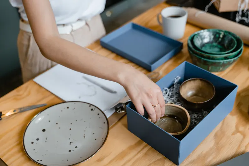 Hands packing handmade bowls into a blue gift box on a workbench.