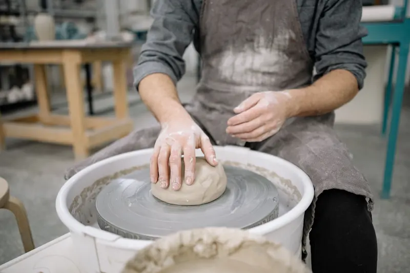 A potter at a wheel in a busy workshop, the kind of equipment a pottery equipment cost calculator prices up.