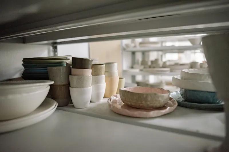 Glazed bowls, cups and plates in soft colours stacked on storage shelves in a pottery studio.