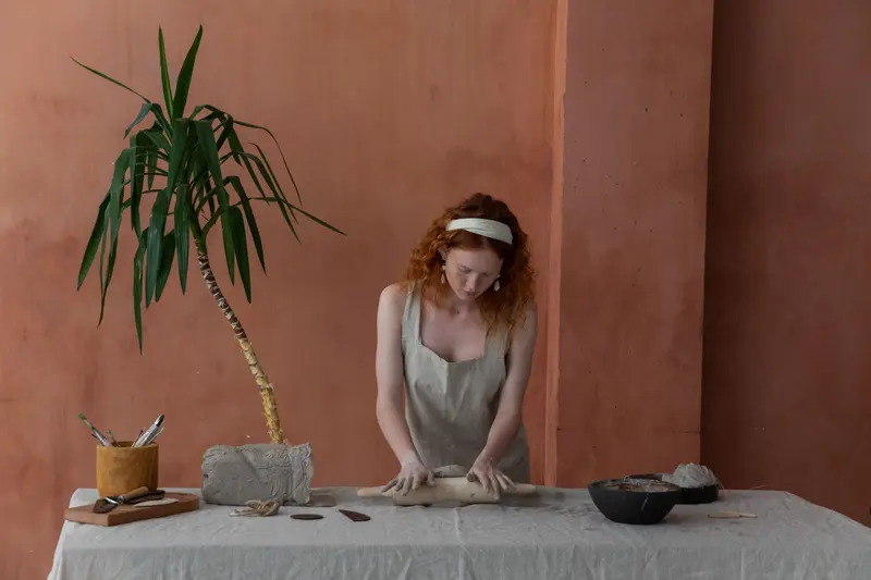A potter in an apron rolling out a clay slab on a long table in a bright studio.
