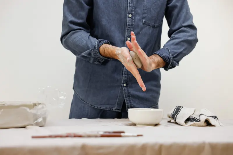 A person in a denim shirt working a small piece of clay between their hands at a studio table.