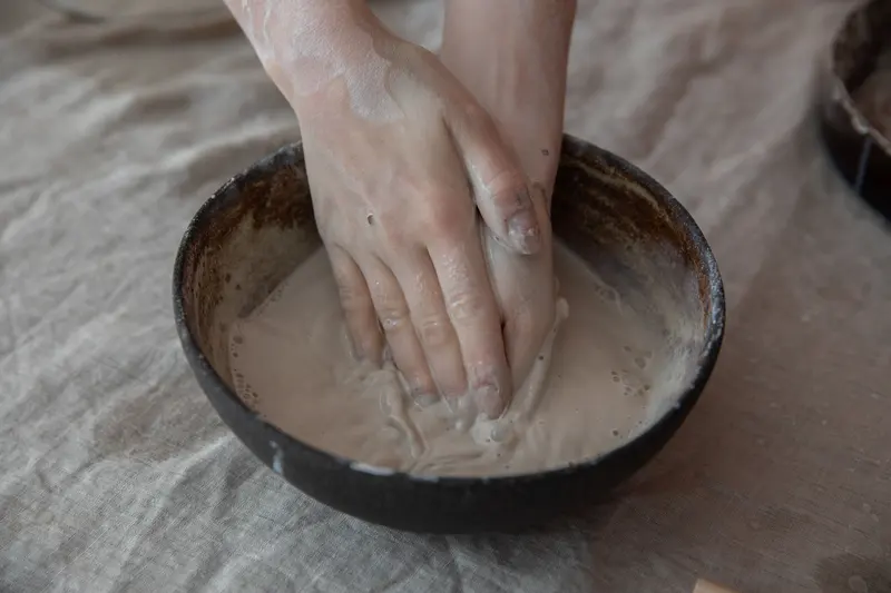 Hands mixing soft clay slop in a round bowl.