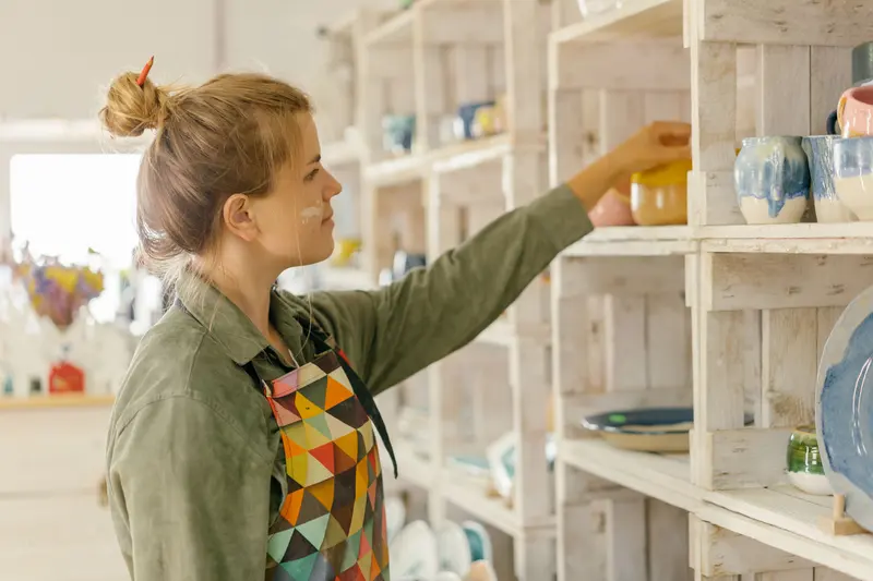 A potter in an apron placing handmade ceramics on white shelves in a bright studio.