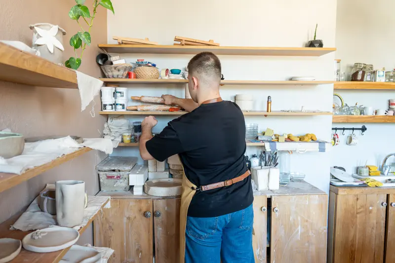 A ceramic artist organising shelves in a pottery workshop, the shelf space that studio hire cost often includes.