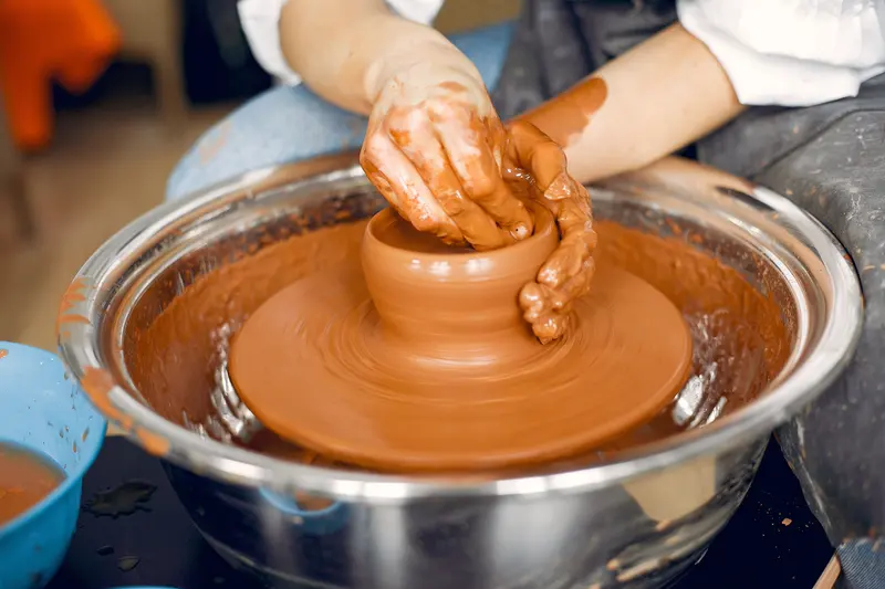 Hands shaping red terracotta clay on a pottery wheel with a splash pan.