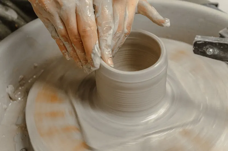 Clay-covered hands pulling up the wall of a cylinder on a spinning wheel head.