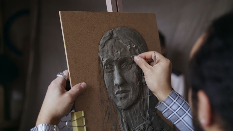 Hands sculpting a clay relief portrait, a form of sculpture that projects from a flat background