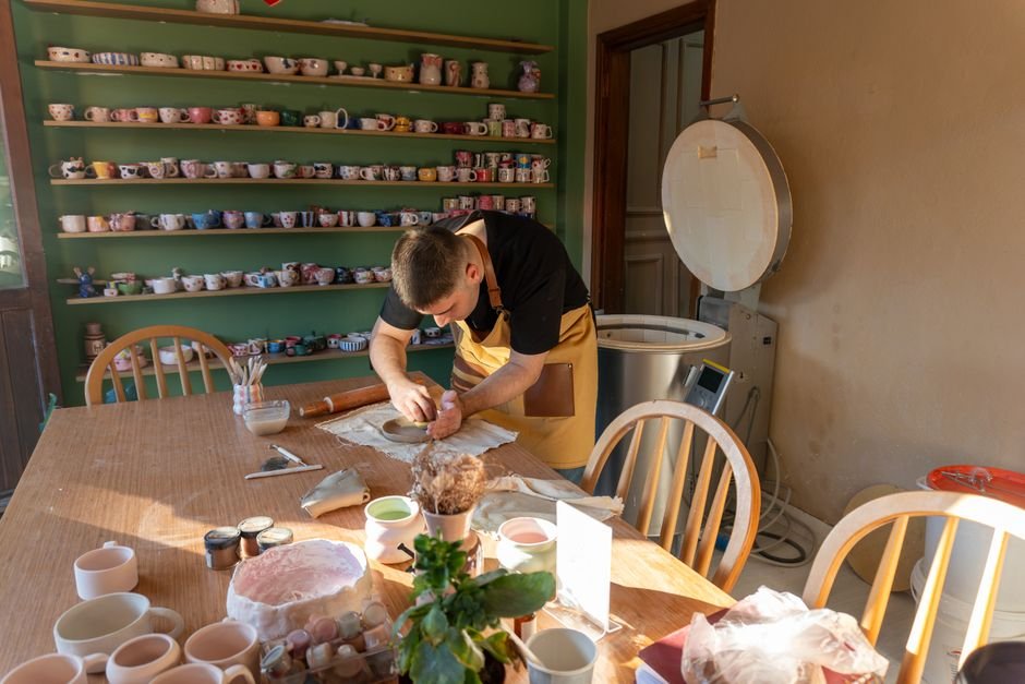 Ceramic artist working in a sunlit studio with shelves of mugs, part of a working ceramic studio layout