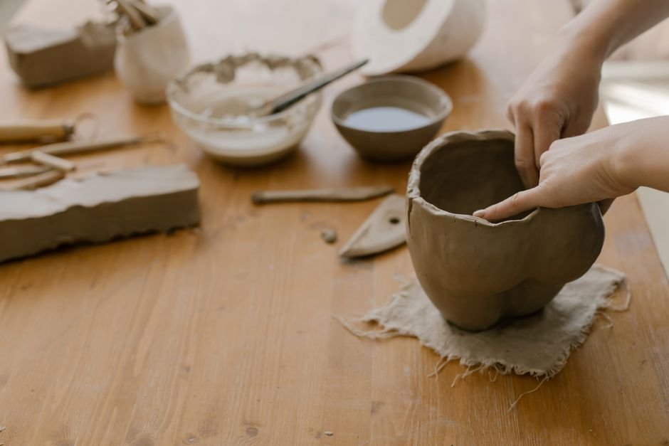 Hands shaping a clay pot on a wooden table, showing the hand building side of wheel throwing vs hand building