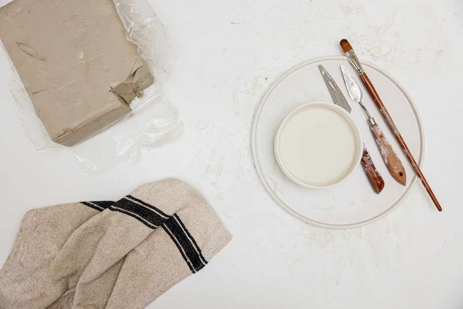 Flat lay of pottery tools and clay on a white background, including blades like a mud knife